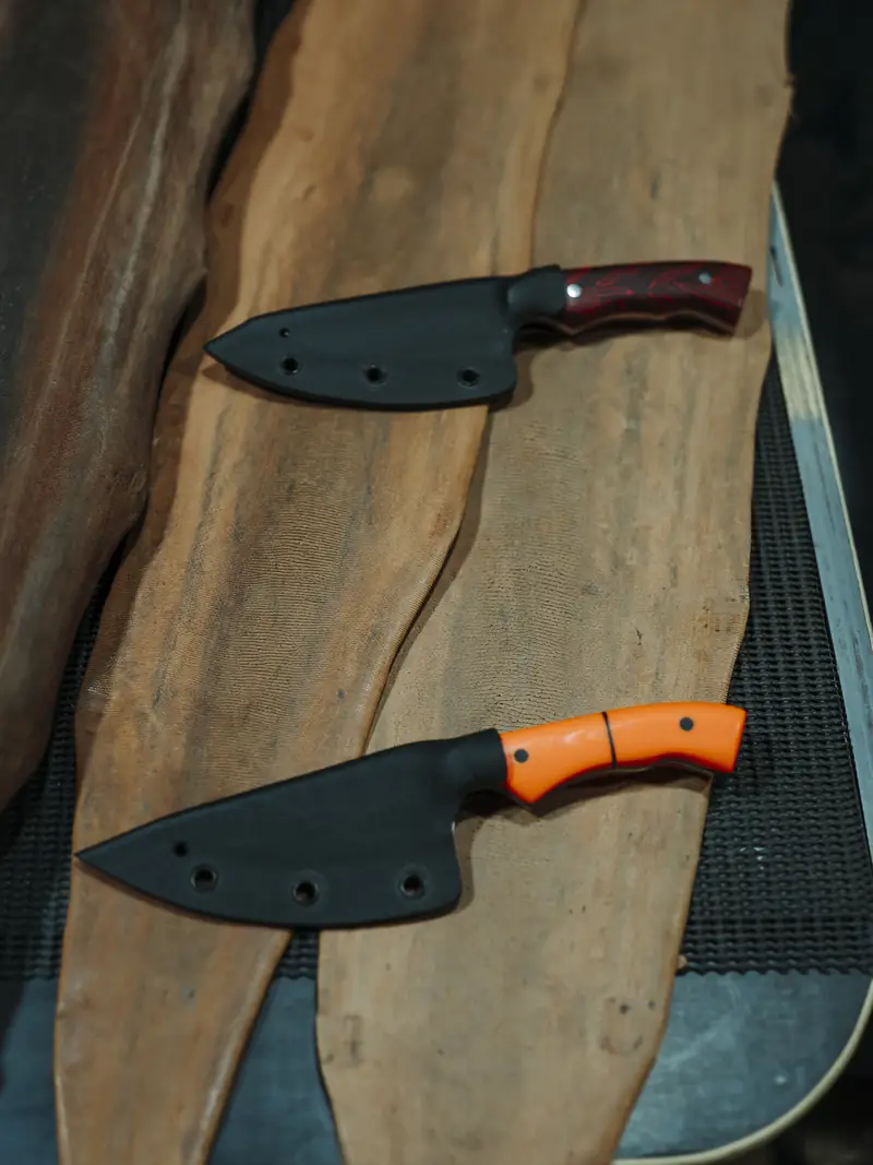 Two custom handmade knives by The Blade Bloke in their kydex sheaths, one with a red handle and one orange